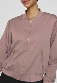 Rosa bomberjakke med stående krave, guld lynlås i lukningen og to sidelommer med lynlås; ribbede manchetter og kant. Blødt, glat stof.