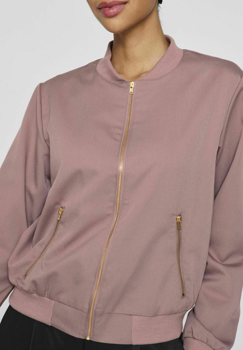Rosa Bomberjacke mit Stehkragen, goldener Reißverschluss und zwei seitlichen Reißverschlusstaschen; gerippte Bündchen und Saum. Weicher, glatter Stoff.