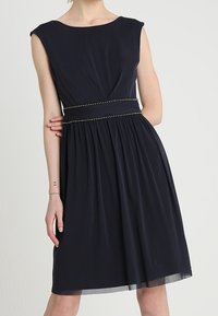 Marineblaues ärmelloses Kleid mit tailliertem Oberteil, plissiertem Rock und dekorativem, mit Nieten verziertem Bund. Hergestellt aus fließendem Stoff mit glatter Textur.
