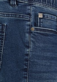 Dark blue denim jeans featuring a front pocket with metal hardware, subtle stitching details, and a branded tag at the pocket edge.