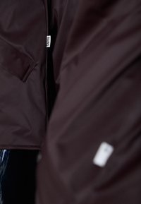 Mørkebrunt vanntett jakke med en glatt tekstur, med en sidelomme og et merke med "RAINS"-logo. Nærbilde av stoffet.