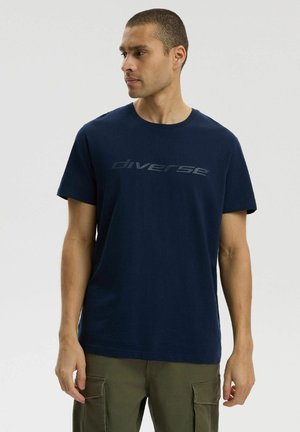 Mężczyzna w granatowej koszulce z napisem "diverse" i oliwkowych spodniach cargo, stoi na tle jednolitego jasnego tła, patrząc w lewo.