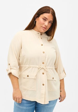 Beige nappi-paita, jossa on naru vyötäröllä, kaksi etutaskua ja käärittävät hihat. Valmistettu pehmeästä kankaasta, jossa on pyöreä kaula-aukko ja puiset napit.