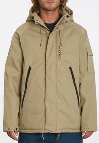 Beige hoodie met een rits aan de voorkant, twee ritszakken en een logo. Bevat verstelbare trekkoorden en een gestructureerde stof.