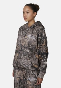 Felpa con cappuccio in stampa camouflage caratterizzata da motivi di foglie marroni, grigie e dorate. Il tessuto appare morbido e presenta una tasca frontale e un dettagli logo.