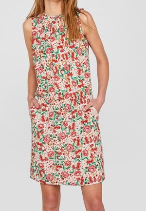 Vestido sin mangas hasta la rodilla con estampado floral en rosa, rojo y verde, llevado por una persona con las manos en los bolsillos sobre un fondo liso.