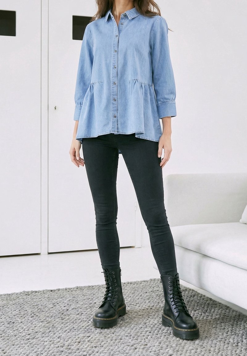 Femme portant un chemisier bleu clair boutonné, un jean skinny noir et des bottes noires à lacets, debout sur un tapis gris à côté d’un canapé blanc.
