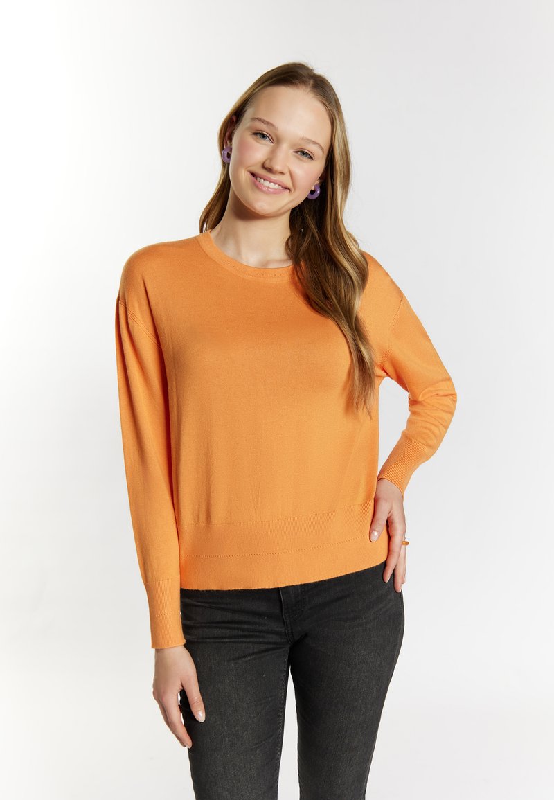 Helles orange gestricktes Sweatshirt mit langen Ärmeln und Rundhalsausschnitt. Weiche Textur. Kombiniert mit schwarzen Jeans, die eine lockere Passform zeigen.