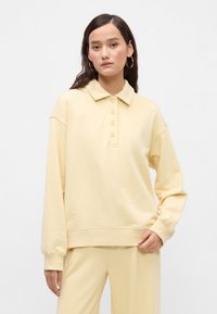 Ανοικτό κίτρινο πολο-τύπου φούτερ  κατασκευασμένο από μαλακό ύφασμα, με τριπλό κουμπί μπροστά, πτώσεις στους ώμους και ριχτές μανσέτες.