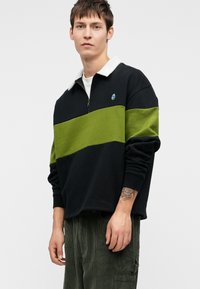 Jaunas vyras, vilkintis juodai žalią raištinę megztinį su balta apykakle ir tamsiai žaliai smulkias velveto kelnes, stovi prieš baltą foną.