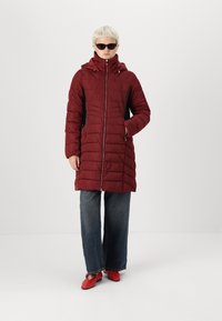 Doudoune rouge avec capuche, dotée d'une fermeture éclair à l'avant, de poches latérales et de panneaux latéraux noirs. Le mannequin porte un jean ample et des chaussures rouges.