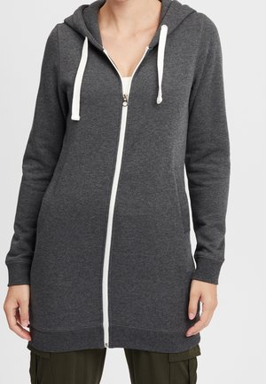 Vrouw draagt een lange antracietgrijze hoodie met rits en witte trekkoorden over een wit topje en olijfgroene broek.