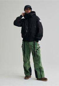 Giacca nera impermeabile con grandi tasche e cerniere, abbinata a pantaloni verdi larghi con dettagli neri e cerniere. Stivali marroni.