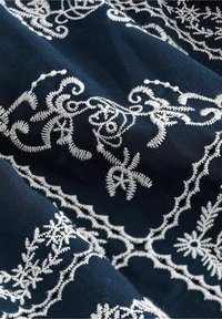 Tissu bleu foncé avec des motifs floraux et arabesques blancs brodés complexes formant des formes rectangulaires et courbes.