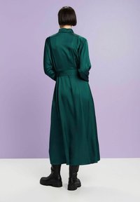 Robe longue en satin vert foncé avec taille nouée, col et manches longues, associée à des bottines noires. Texture lisse et design fluide.