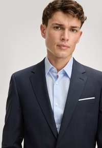 Granatowa marynarka o drobnej fakturze i klapach, występująca w zestawie z jasnoniebieską koszulą z delikatnym wzorem i białymi guzikami.