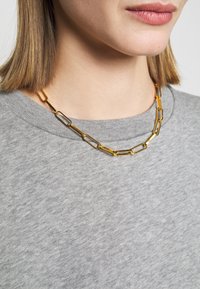 Primer plano de una persona que lleva un collar de cadena con eslabones rectangulares dorados y un suéter gris de cuello redondo.