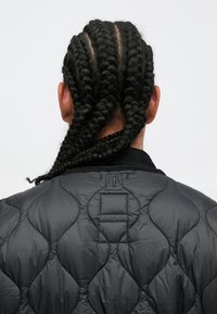 Chaqueta acolchada negra con una capa exterior sin costuras, cuello alto, y trenzas texturizadas en el cabello en la parte posterior. Diseño minimalista sin marcas visibles.
