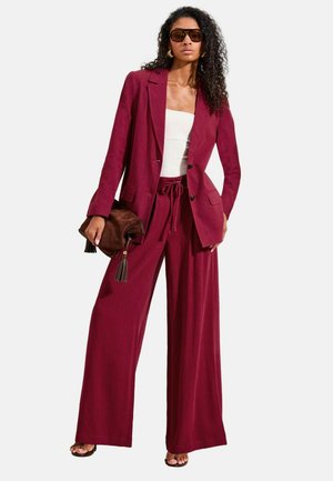 Femme portant un pantalon bordeaux à jambes larges et un blazer, un top tube blanc, des lunettes de soleil foncées, tenant une pochette à franges marron, posant devant un fond blanc.
