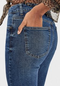 Jeans de denim con un lavado azul medio, con un bolsillo trasero y detalles de costura visibles. La tela tiene una apariencia suave y ligeramente texturizada.
