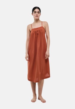 Femme debout pieds nus, portant une robe midi sans manches de couleur rouille avec de fines bretelles et un tissu froncé sous le décolleté.