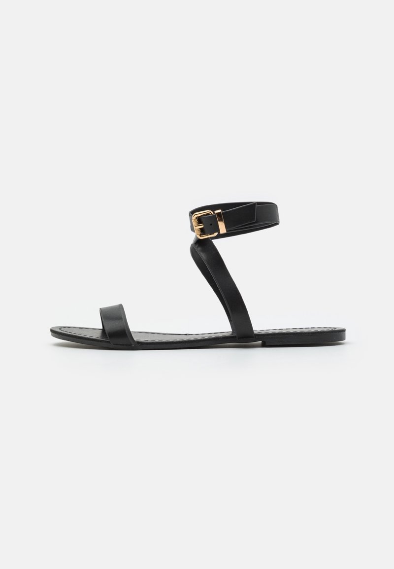 Sandale noire en cuir avec deux fines lanières et une lanière de cheville avec une boucle en or. Semelle plate, design minimaliste, texture lisse.