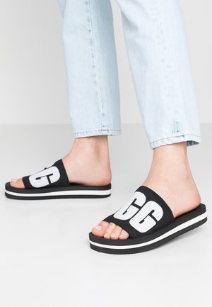 Füße in hellblauen Jeans und schwarzen Plateau-Pantoletten mit weißen "GG"-Logos auf den Riemen vor neutralem Hintergrund.