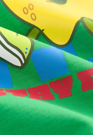 Zielona tkanina z wzorami w żółte i niebieskie kształty, nałożony jest wyrazisty czerwony tekst. Materiał wydaje się mieć fakturę z widocznym przeszyciem.