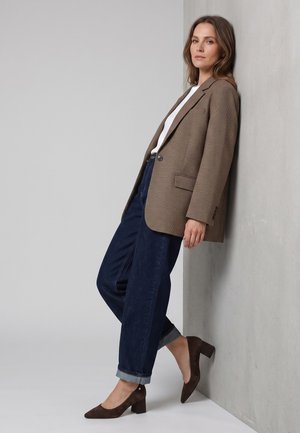 Frau trägt braun karierten Blazer, weißes Top, dunkelblaue hochgekrempelte Jeans und braune Schuhe mit Blockabsatz und lehnt an einer grauen Wand.