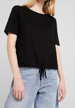 Mujer que lleva una camiseta negra texturizada de manga corta con cordón en el dobladillo, combinada con unos vaqueros de tiro alto en azul claro sobre un fondo liso.