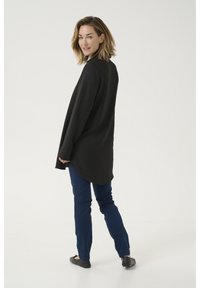 Schwarzes Langarmshirt mit strukturierter Oberfläche, abgerundetem Saum, kombiniert mit blauen Slim-Fit-Jeans und schwarzen Ballerinas, von hinten gezeigt.