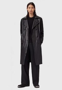 Person med långt hår bär en lång svart lädertrenchcoat, svart tröja, vida svarta byxor och svarta skor mot en neutral bakgrund.