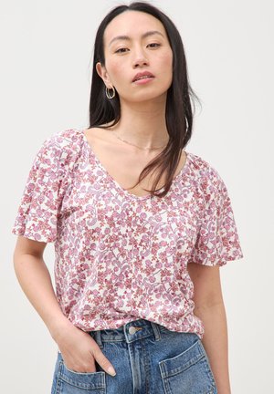 Vrouw met lang donker haar, gouden oorringen, een roze blouse met korte mouwen en bloemenprint, en een blauwe spijkerbroek met één hand in de zak.