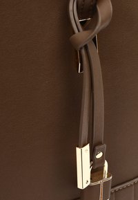 Close-up of brown leather bag with thin straps and gold-toned metal hardware detail and stitching along the edge.