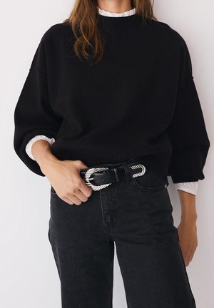 Frau trägt schwarzen übergroßen Pullover über weißer Bluse mit gerafften Manschetten, hoch taillierte schwarze Jeans und einen schwarzen Gürtel mit silberner, verdrehter Schnalle.