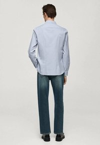 Mies, který je viděn zezadu, má na sobě světlemodrou košili s dlouhým rukávem, rovné modré džíny a černé boty, stojící na jednobarevném pozadí.