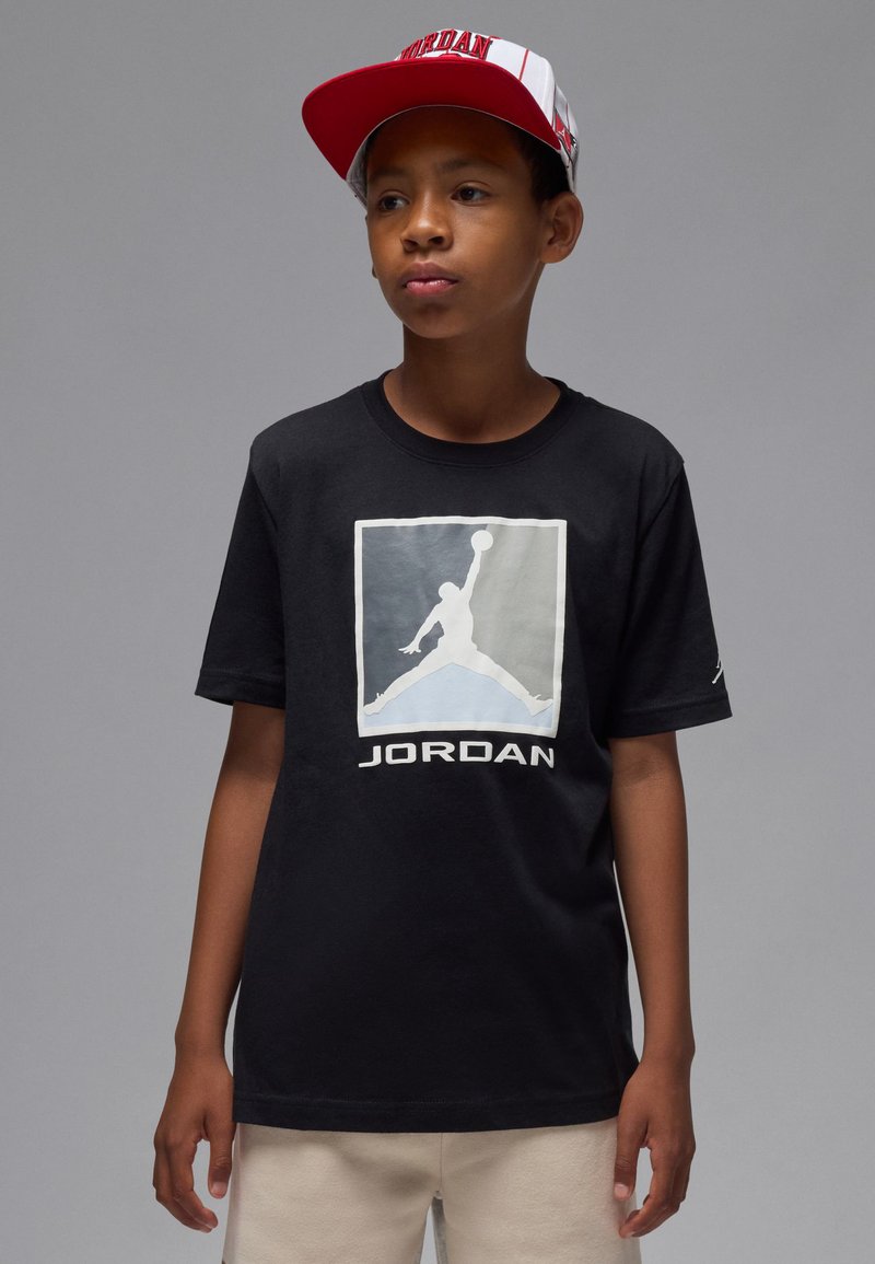 T-shirt en coton noir arborant un logo Jordan blanc et gris bien visible. Associé à une casquette de baseball rouge et blanche avec le texte « Jordan ».