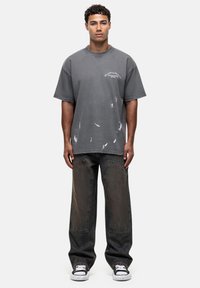 Szary, oversize T-shirt z białymi przetarciami, w zestawieniu z luźnymi, ciemnymi dżinsami. Model nosi czarno-białe trampki. Prosty design.