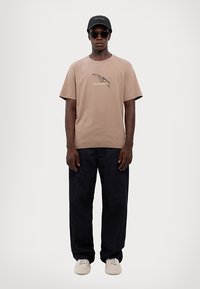 Beige Baumwoll-T-Shirt mit grafischem Druck, kombiniert mit dunklen Denim-Hosen. Mit schwarzer Cap und Sonnenbrille accessoriert; die Schuhe sind weiße Slipper.