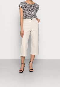 Mujer vestida con una blusa de manga corta de estampado animal en blanco y negro, pantalones cortos de pierna ancha en crema de cintura alta y tacones negros con tiras al tobillo.