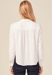 Weißes Langarmhemd mit einem strukturierten Muster, gefälteter Rückseite, klassischem Kragen und geknöpften Manschetten, kombiniert mit dunklen Jeans.