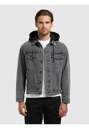 Mann mit kurzen braunen Haaren, trägt eine graue Jeansjacke über einem schwarzen Kapuzenpullover, einem weißen T-Shirt und schwarzen Hosen.