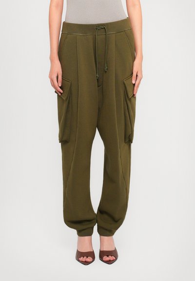 Femme portant un pantalon cargo jogger vert olive avec taille à cordon et des talons ouverts bruns, se tenant devant un fond uni.