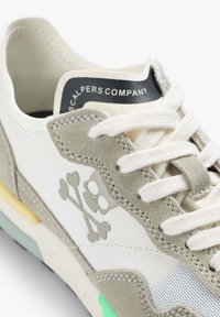 Sneaker w białym materiale i szarym zamszu z logo czaszki i skrzyżowanych kości. Posiada teksturowane akcenty, jasnooliwkowe i żółte detale oraz białe sznurówki.