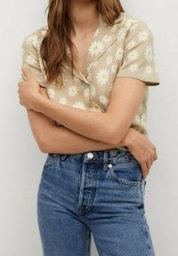 Beżowa koszula z krótkim rękawem z białymi i żółtymi wzorami stokrotek, zestawiona z niebieskimi jeansami z wysokim stanem. Gładki materiał i dopasowany krój.