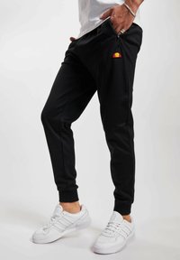 Pantalones de chándal negros con bolsillos con cremallera, presentando un logo colorido en la cadera izquierda. Combinados con zapatillas deportivas blancas.