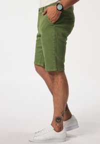 Zelené bavlněné šortky se slim fit, dvěma bočními kapsami a dvojitým zapínáním na knoflíky, v kombinaci s bílými teniskami a tmavými hodinkami.