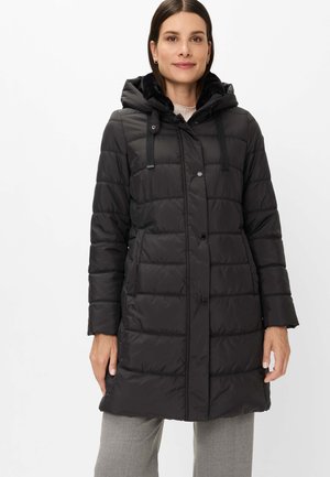 Woman wearing a knee-length black quilted coat with a hood, button and zipper closure, and light gray pants, standing against a white background.