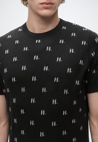 Jeune personne portant un t-shirt noir à manches courtes avec des motifs répétés de petits logos blancs HL et un col rond.