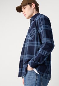 Camisa de flanela xadrez azul-marinho com padrões de xadrez azul-claro, frente com botões e bolso no peito. Usada com jeans de ganga clara.